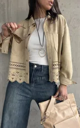Sobrecamisa beige de gamuza con cuello camisero, cierre frontal con botones y mangas largas con puños abotonados. Presenta bolsillos de parche en el pecho y detalle de calado en el ruedo.