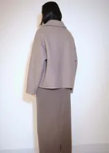 Chaqueta color gris topo de tejido bouclé, con cuello camisero, cierre frontal con botones, dos bolsillos de parche con solapa y botón en el pecho, y mangas largas.