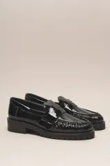 Mocasín negro de cuero vacuno con acabado charol, suela de goma y forro y plantilla de cuero vacuno.