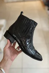 Bota corta de cuero vacuno color negro charol, con elástico lateral, punta fina y detalles de calado.