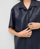 Camisa de manga corta, color azul marino, confeccionada en material que simula cuero (piel no piel). Presenta cuello solapa, botonadura frontal y un bolsillo de parche en el pecho.