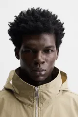 Parka color beige de tejido técnico ligero, corte relaxed fit, cuello alto con capucha plegable, manga larga con puños ajustables con botón a presión, cierre frontal con cremallera, cintura ajustable con elástico interno y múltiples bolsillos frontales de parche con solapa.
