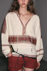 Blusa color crema de cuello redondo con escote pico y lazadas. Manga larga abullonada. Detalle de bordados combinados a contraste en cuello, mangas, pecho y puños. Cierre frontal con botones.