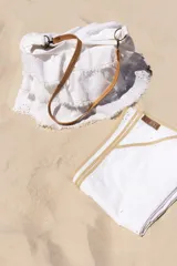 Bolso blanco de lino bordado con volados superpuestos y desflecados a mano. Está forrado con gabardina de algodón marfil y tiene bolsillo interno de lino. Se cuelga con correa de cuero color camel que se puede sacar ya que viene con mosquetones como herraje que se enganchan en argollas a los costados. El bolso se cierra con broche interno de imán.