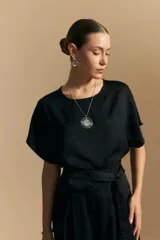 Top negro de manga corta con silueta amplia y caída fluida, confeccionado en un tejido liviano con leve brillo. Presenta un botón de nácar en la espalda con una pequeña abertura y mangas amplias tipo kimono.