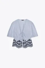 Blusa de rayas verticales azules y blancas, con escote en pico, mangas cortas abullonadas con elástico y bajo con bordados florales perforados. Cierre frontal con lazos.