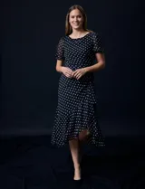 Vestido midi de gasa color negro con lunares blancos, con mangas cortas, escote redondo y ruedo asimétrico. Presenta un frunce lateral con lazo de ajuste.