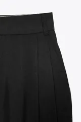 Pantalón negro de tiro alto con trabillas, doble pliegue, bolsillos laterales y tapetas en la espalda. Tiene pernera ancha y cierre frontal con cremallera y gancho metálico.