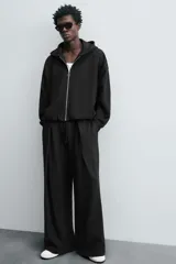 Pantalón jogger negro de corte relaxed fit, confeccionado en tejido de algodón. Presenta cintura elástica ajustable con cordón, pliegues frontales, bolsillos delanteros y un bolsillo trasero.
