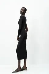 Vestido midi negro de punto, con cuello asimétrico, manga sisa, aplique metálico dorado en el hombro y drapeado lateral.