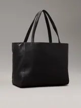 Cartera tote Calvin Klein color negro, confeccionada en piel sintética, con doble asa de hombro, cierre superior con cremallera, bolsillo externo con cierre giratorio y logo CK plateado.