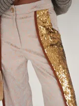 Pantalón de brocato color champagne con lentejuelas doradas en los laterales, tiro medio, cuatro bolsillos, cierre frontal con botón de guampa y bajo acampanado con tajos laterales.