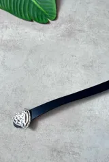 Cinturón negro de cuero sintético con hebilla metálica plateada en forma de corazón martillado y borde con pequeñas incrustaciones brillantes.