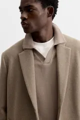Abrigo recto color beige confeccionado en tejido con lana. Tiene cuello con solapas de muesca, manga larga con puños abotonados, bolsillos de vivo en la cadera, bolsillo interior, abertura en el centro de la espalda y cierre frontal con botonadura.