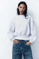 Pantalón de jean celeste de tiro alto, corte wide leg y largo hasta el tobillo. Presenta un diseño asimétrico en la cintura con cierre frontal cruzado con cremallera y botones metálicos.