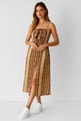 Vestido midi de tirantes finos, color marrón con estampado de hojas en beige. Tiene escote recto con detalle de lazo y abertura lateral en la falda.