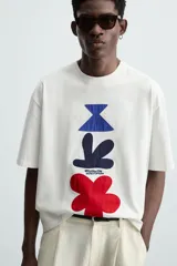 Camiseta blanca de corte regular, cuello redondo y manga corta. Presenta parches geométricos en azul, negro y rojo en el frente y estampado en la espalda.