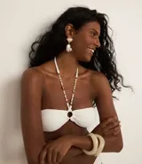 Top de bikini bandeau blanco con textura acanalada e hilo metalizado. Tiene aro en el busto, copas removibles y lazo en el cuello con adornos de caracolas y perlas artificiales.