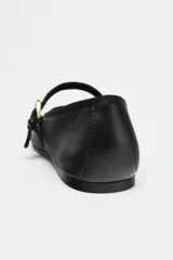 Zapato plano tipo bailarina color negro, confeccionado en piel, con tira delantera con hebilla metálica ajustable y acabado en punta redonda.
