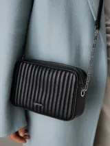 Cartera bandolera Calvin Klein negra con diseño acolchado de líneas verticales, correa de cadena plateada, cierre superior con cremallera y logo metálico frontal.