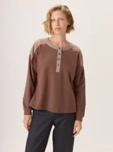 Buzo de manga larga con diseño de bloques de color y textura. El cuerpo principal es marrón, la espalda superior y hombros tienen un panel de tejido a rayas grises y azules, y los hombros superiores son de color beige rosado. Presenta costuras decorativas en rojo que separan los bloques de color.