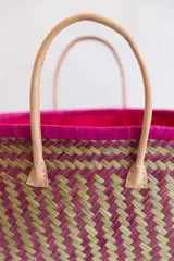Bolso tipo tote de raffia natural con diseño tejido, interior forrado en tela fucsia y doble asa de cuero color beige. Tiene base rectangular y detalles de cuero en las esquinas inferiores.