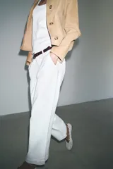 Pantalón cropped color beige de tiro alto y corte recto, confeccionado en algodón. Cuenta con bolsillos delanteros y traseros, trabillas en la cintura y cierre frontal con cremallera y botón.