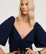 Vestido corto de viscosa con estampado floral y de follaje en tonos mostaza, blanco y azul marino. Presenta escote pronunciado en V con lazo y fruncido en el busto, mangas cortas abullonadas y elástico (lastex) en la espalda. La falda es evasé con un ruedo con volado.