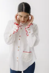 Camisa blanca de lino con cuello mao, manga larga abullonada y puños con detalle de crochet rojo. Tiene bolsillo de crochet en el pecho y cierre frontal con botones de madera.