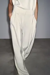 Pantalón fluido color crudo, de tiro medio con cintura con trabillas, bolsillos laterales, pinzas y raya planchada en delantero. Cierre frontal con cremallera, botón y ganchos metálicos.