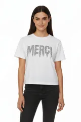 Remera blanca de algodón con cuello redondo y manga corta. Estampado frontal con la palabra "MERCI" en negro con efecto de puntos o tachas.