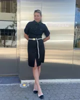 Vestido camisero negro de lino con cinturón de tela a rayas blancas y negras con hebilla dorada. Tiene cuello camisero, cierre frontal con botones, mangas cortas con trabillas y bolsillos en el pecho y laterales.