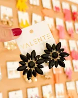 Aretes tipo colgante con diseño de flor grande, compuestos por múltiples piedras de cristal facetadas de color negro dispuestas en pétalos alrededor de un cristal central redondo. La base es de metal dorado.