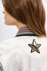Campera bomber de cuero ovino color plateado, con cuello, puños y ruedo de ribb metalizado. Presenta apliques de parches bordados con lentejuelas y mostacillas.