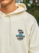 Hoodie color crema con capucha y manga larga, de corte holgado y estampado frontal con un personaje de caricatura y la frase "Totally Legit".