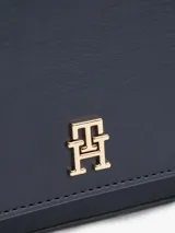 Cartera pequeña de poliuretano color azul marino con acabado liso. Presenta un diseño rectangular con solapa frontal que se cierra magnéticamente y lleva el monograma TH en metal dorado al frente. Incluye correa ajustable y extraíble para llevar al hombro o cruzada.