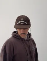 Gorro tipo gorra de béisbol color marrón, confeccionado en pana, con visera corta y bordado frontal en blanco que dice "LOVE & PEACE" y "PP:ONE".