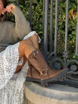 Bota de caña alta color marrón chocolate, realizada en cuero encerado. Presenta hebillas en la caña, herrajes de níquel y cierre lateral con cremallera. El interior está forrado en cuero vacuno y el fondo es de suela color negro.