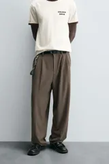 Pantalón negro de corte ancho con cintura elástica, pliegues frontales, bolsillos laterales y traseros de vivo. Tiene detalle de hebilla metálica en la trabilla y cierre frontal con cremallera y botón.