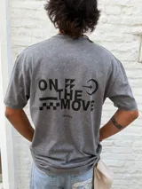 Remera gris con efecto acid wash, corte oversize y cuello redondo. Presenta un pequeño estampado negro con el texto "ON THE MOVE" y un gráfico de damero en el pecho.