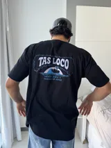 Remera negra de corte holgado con estampado gráfico en la espalda que dice "TAS LOCO" en letras rosas y celestes, con una ilustración de una ola y el texto "SUNNY WITH A CHANCE OF STOKE".