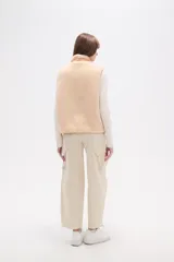 Chaleco holgado confeccionado en poliéster y corderito en tonos beige y crema. Presenta cuello alto, cierre frontal con botones a presión y un bolsillo con cierre en el pecho. Combina texturas de corderito con paneles de tela lisa.