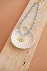 Collar corto con cuentas de cristal celestes y blancas, y dije dorado con forma de sol y circonitas.