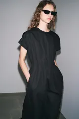 Vestido midi color marrón confeccionado en hilatura con mezcla de algodón y lino. Tiene cuello redondo, manga corta caída y detalle de pliegues en la cintura. Cierre en la espalda con abertura y botón.