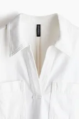 Vestido corto blanco de denim rígido, corte recto y holgado, con cuello camisero, escote en V, mangas cortas y bolsillos de parche en el pecho.
