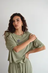 Remera de manga corta color crema, con textura acanalada y cuello redondo. Presenta un pequeño parche bordado con un diseño floral o geométrico en el pecho.