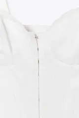 Top blanco estilo corsé con escote corazón, tirantes finos y bordados a tono. Presenta un bajo con volantes y cierre frontal con ganchos metálicos. La espalda es de tejido elástico tipo nido de abeja.