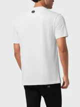 Camiseta blanca de manga corta con cuello redondo. Estampado frontal con la palabra "PLEIN" en tipografía gótica negra, flanqueada por dos líneas horizontales y un emblema central con detalles florales y texto pequeño.