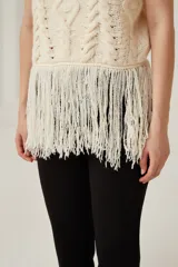 Chaleco de punto sin mangas color beige, con cuello alto tipo polera. Presenta un diseño de tejido con ochos y rombos, y terminación de flecos largos en el ruedo.