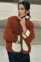 Campera marrón de tela tipo sherpa con cuello, puños y bolsillos de tela blanca. Tiene cierre con botones a presión y ribete azul.